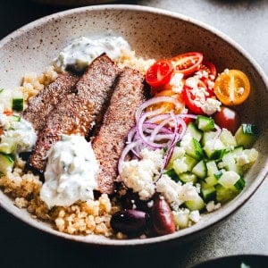 A bowl filled with quinoa, sliced gyro meat, diced cucumbers, cherry tomatoes, red onion slices, olives, crumbled feta cheese, and dollops of tzatziki sauce. The dish is colorful and arranged in a visually appealing way.