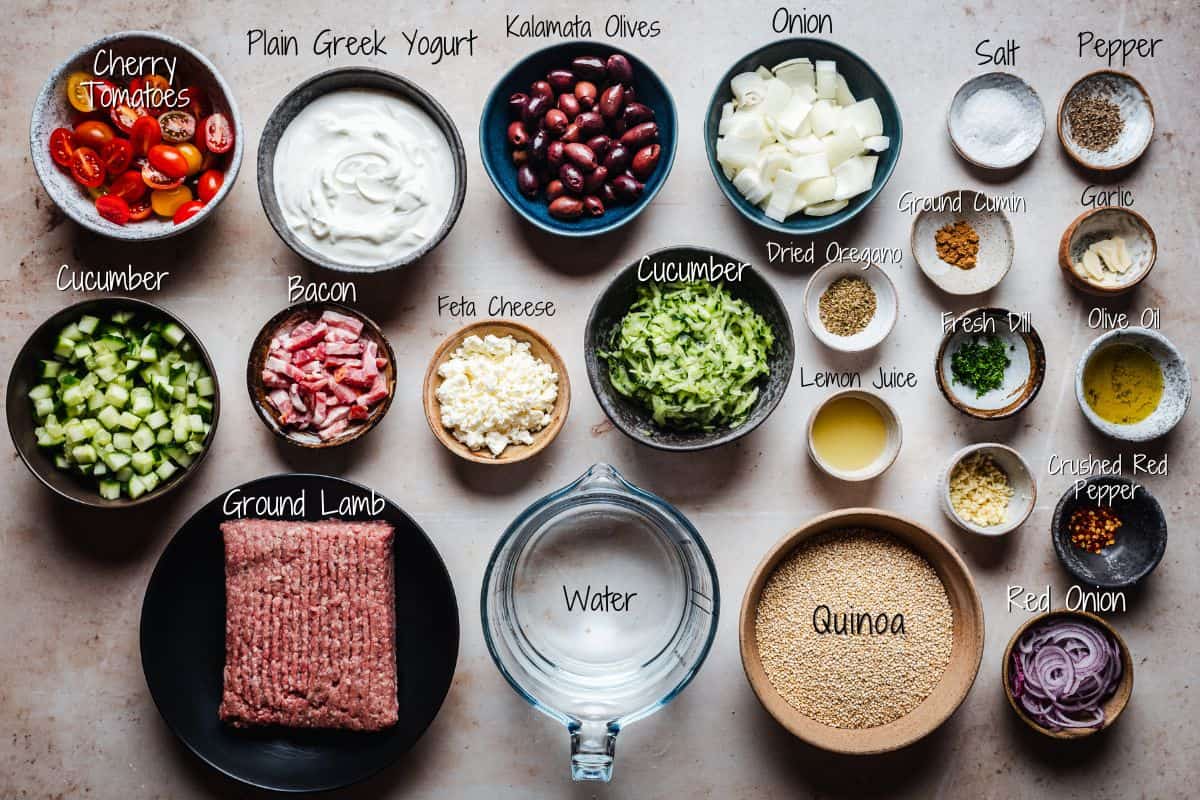 Top-down view of Gyro Bowl ingredients arranged on a surface, including ground lamb, quinoa, bacon, Greek yogurt, cherry tomatoes, cucumbers, Kalamata olives, feta, onion, water, oil, and various spices labeled with handwritten text.
