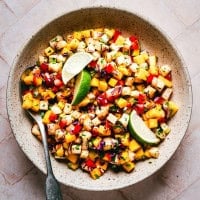 A bowl of colorful fruit salsa featuring diced pineapple, mango, and red bell peppers, garnished with chopped herbs and lime wedges. A metal spoon rests inside the bowl on a light tiled surface.