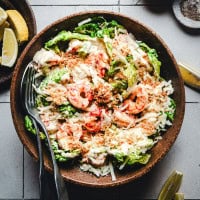 A wooden bowl filled with a Caesar salad topped with grilled shrimp, shredded cheese, and croutons. A fork and spoon rest on the side. Lemon wedges are in a dish nearby on a tiled surface.