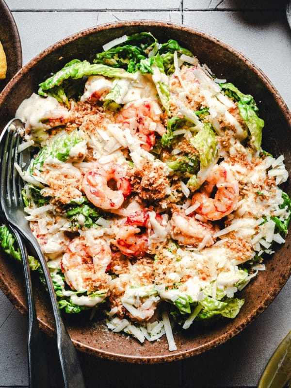 A wooden bowl filled with a Caesar salad topped with grilled shrimp, shredded cheese, and croutons. A fork and spoon rest on the side. Lemon wedges are in a dish nearby on a tiled surface.