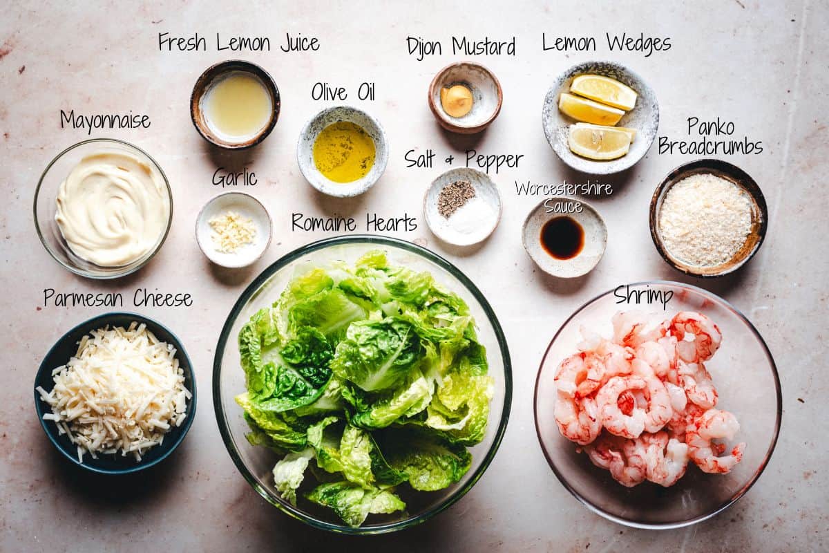 Shrimp Caesar Salad Ingredients on a table: mayonnaise, Parmesan cheese, fresh lemon juice, olive oil, romaine hearts, salt and pepper, garlic, Dijon mustard, Worcestershire sauce, lemon wedges, panko breadcrumbs, and shrimp.