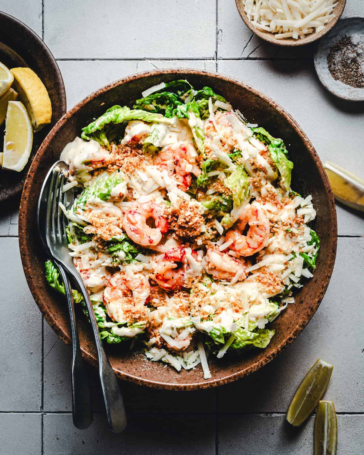 A bowl of Caesar salad topped with grilled shrimp, shaved parmesan, croutons, and creamy dressing. Lemon wedges and a bowl of grated cheese are on the side, with a fork and spoon placed inside the bowl.