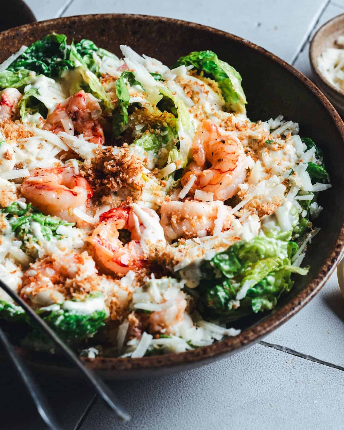 A salad in a brown bowl featuring grilled shrimp, shredded cheese, and mixed greens, topped with breadcrumbs. A fork is placed on the side. The table is tiled and sunlight illuminates the dish.