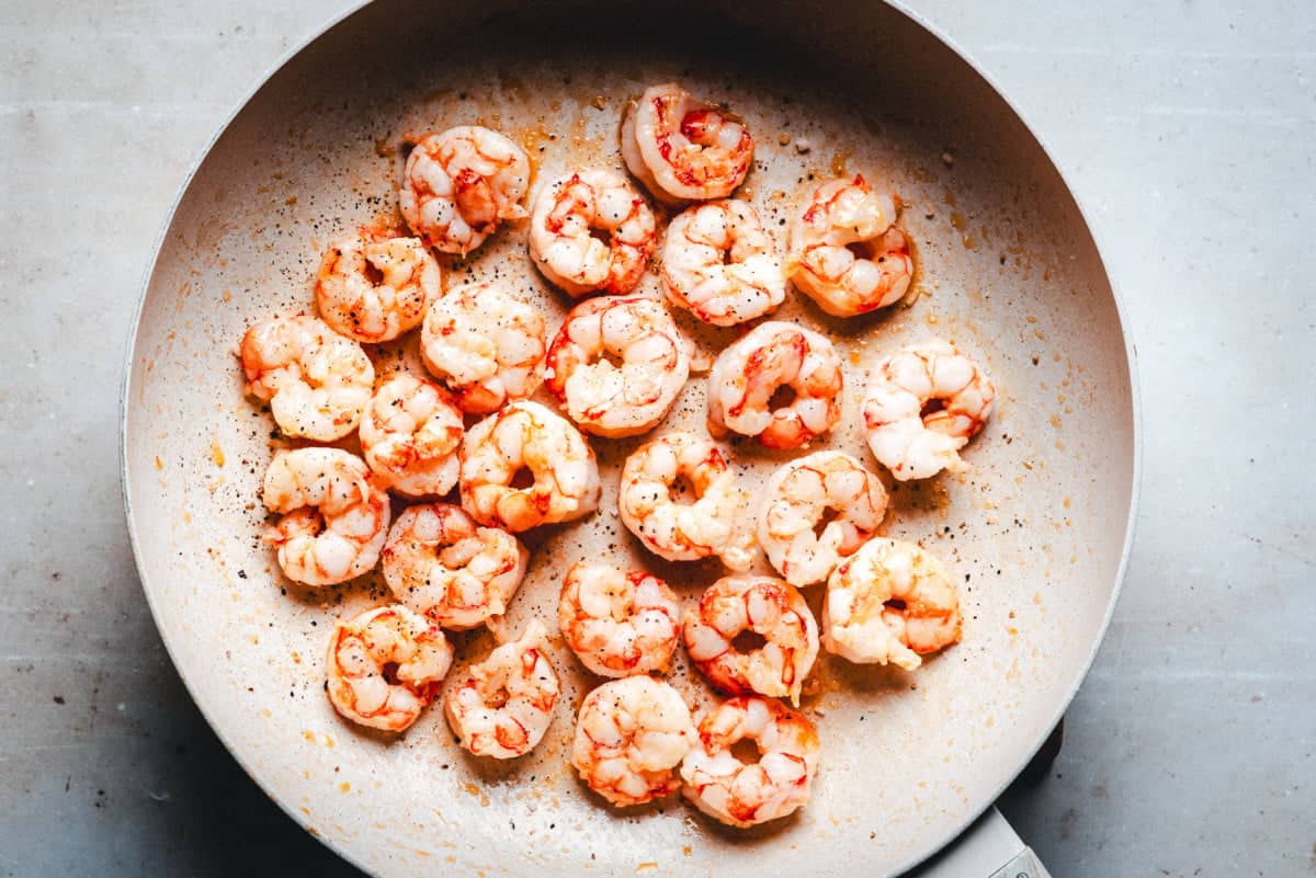 A top view of a frying pan filled with cooked shrimp. The shrimp are seasoned with herbs and spices, giving them a slightly reddish hue. The pan is on a light gray surface.