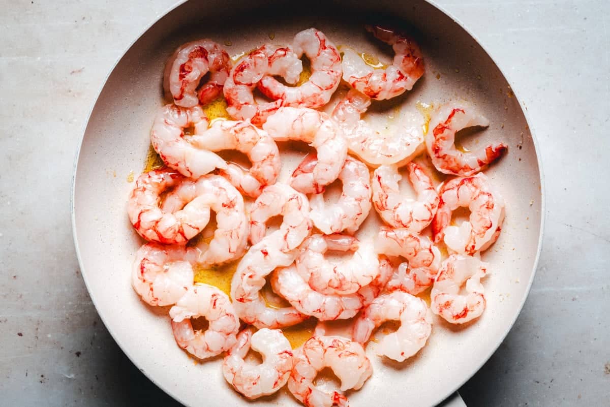 A frying pan filled with raw pink shrimp in a light coating of oil, placed on a gray surface. The shrimp are arranged evenly, with visible marbling of pink and white colors.