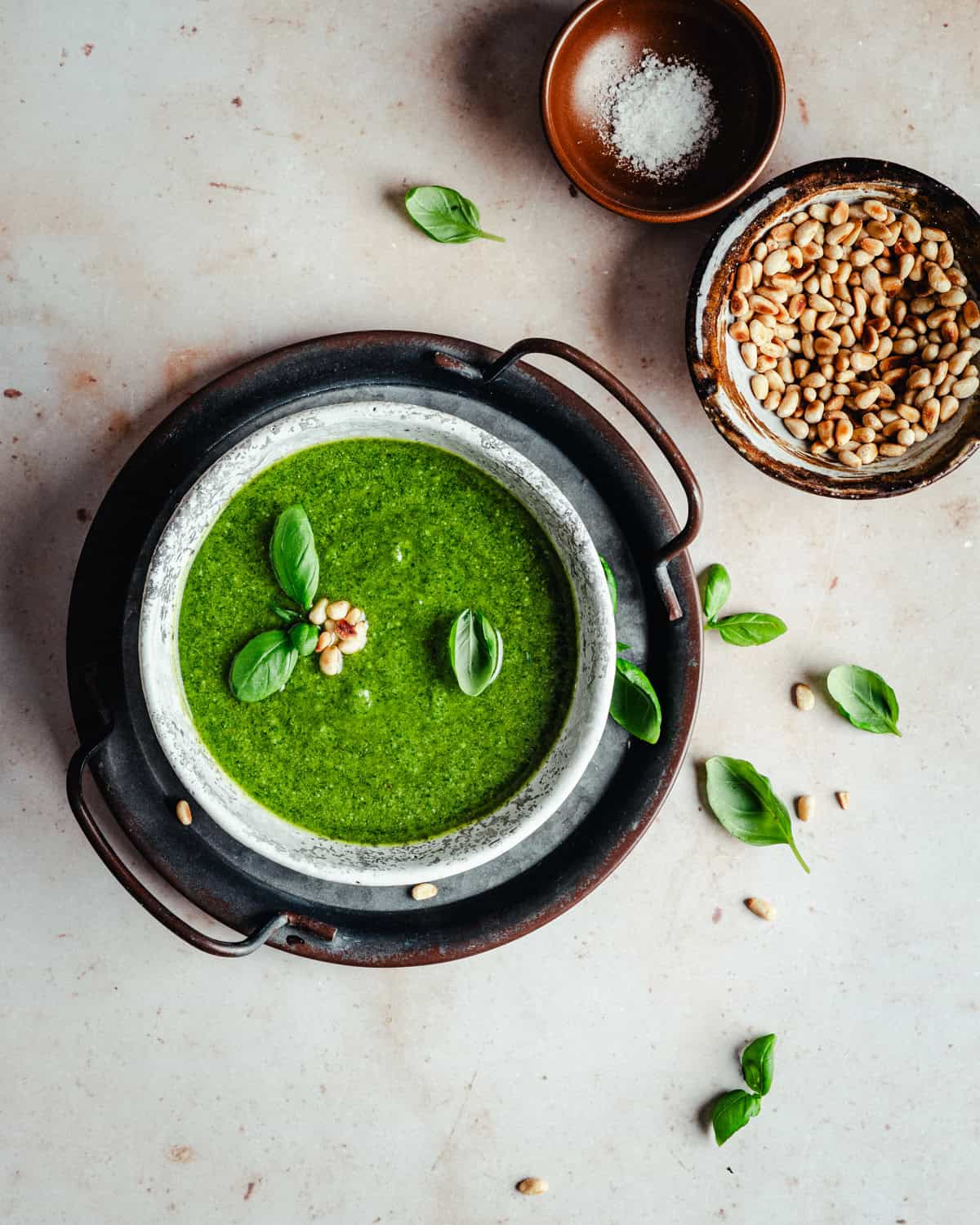A bowl of green pesto garnished with basil leaves and nuts, placed in a dark tray. Next to it are bowls containing salt and pine nuts, with scattered basil leaves and nuts on the light surface.
