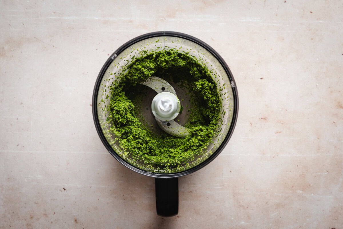 Top view of a food processor containing freshly processed pesto, against a light marble surface. The metal blade is visible in the center.