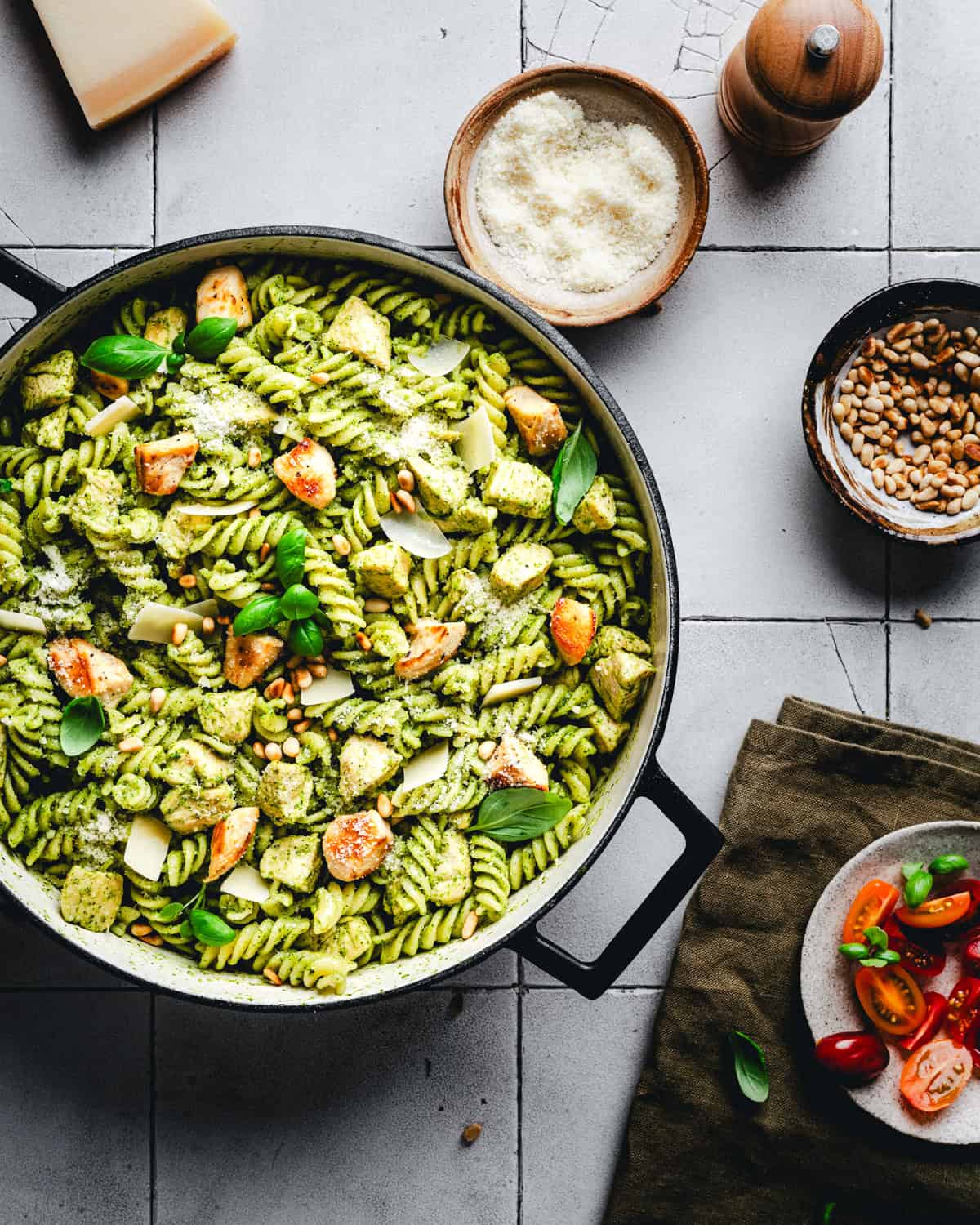 A skillet filled with pesto pasta topped with chicken, pine nuts, and fresh basil. Surrounding it are bowls of grated cheese, pine nuts, and a dish with sliced tomatoes. A pepper grinder is also visible on a light tiled surface.