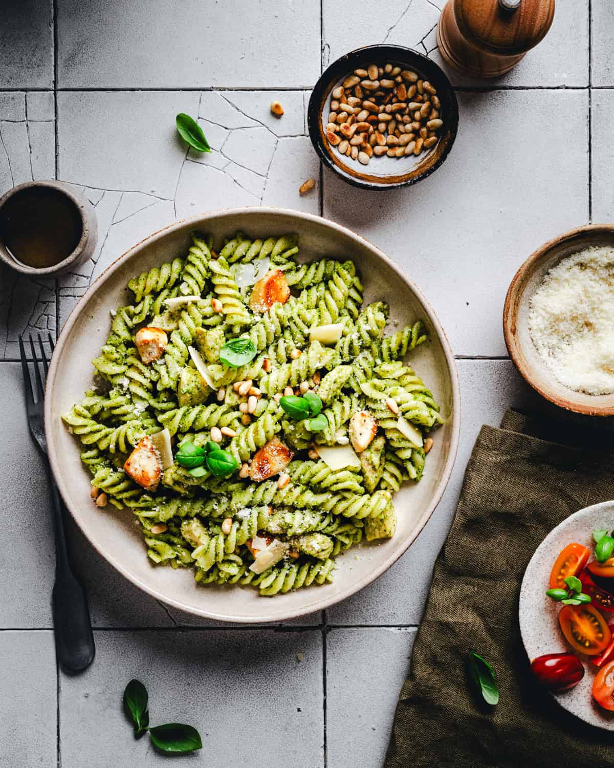 A bowl of pasta with pesto sauce, chicken, pine nuts, and basil is placed on a tiled table. Nearby are bowls of grated cheese and pine nuts, a pepper grinder, a cup, and scattered cherry tomatoes and basil leaves.
