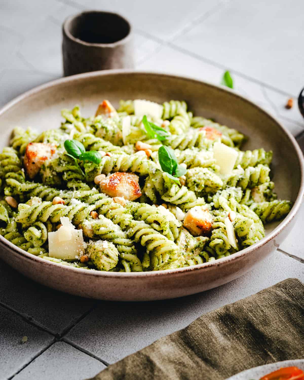 A bowl of fusilli pasta coated in green pesto sauce, topped with chunks of chicken, parmesan cheese shavings, pine nuts, and basil leaves. The bowl is set on a tiled surface with a small cup in the background.