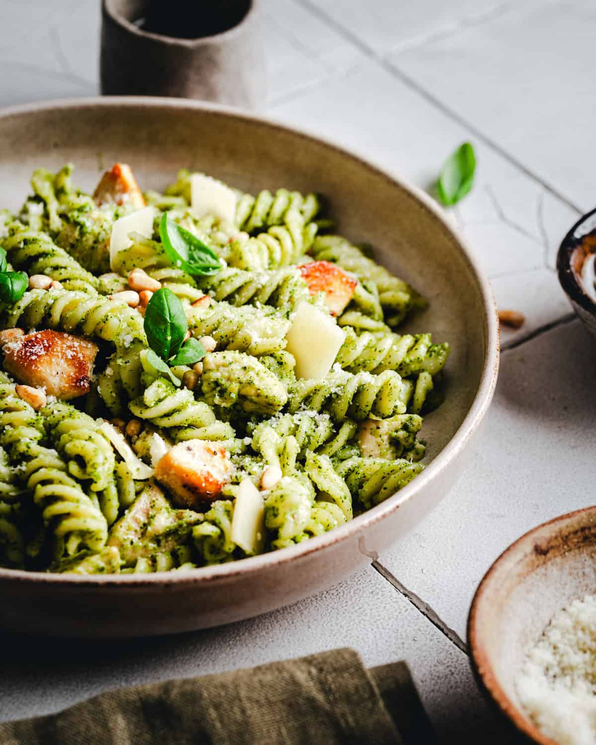 A bowl of fusilli pasta coated in green pesto sauce, garnished with chicken pieces, parmesan shavings, and fresh basil leaves. The dish is set on a light-colored surface with additional bowls in the background.