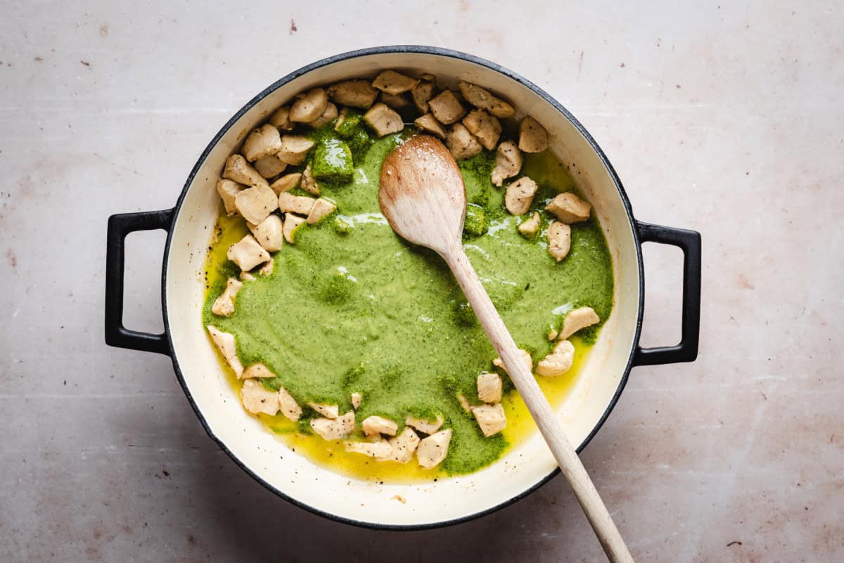 A pot with cubed chicken pieces and a vibrant green creamy pesto sauce, stirred with a wooden spoon. The pot has black handles and is placed on a light-colored surface.