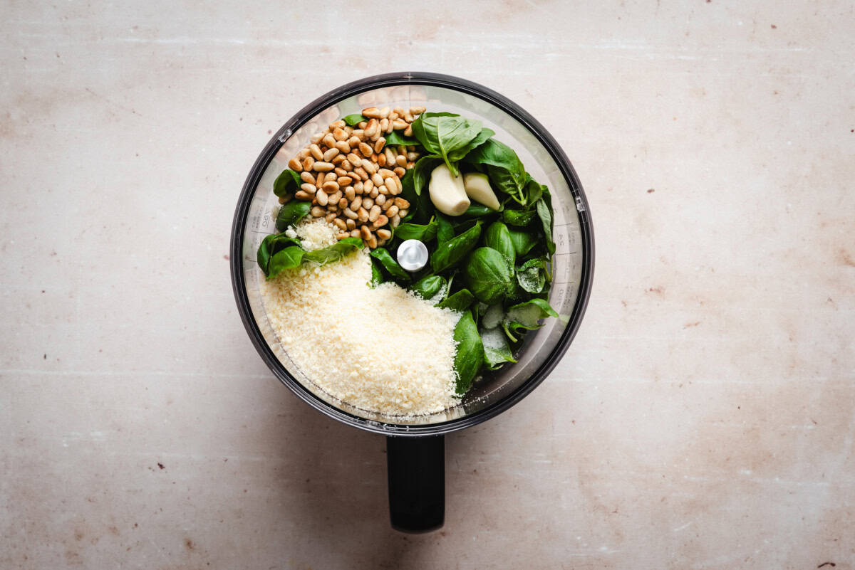 Food processor bowl with fresh basil leaves, grated Parmesan cheese, pine nuts, and a garlic clove, set on a light countertop, ready for making pesto.