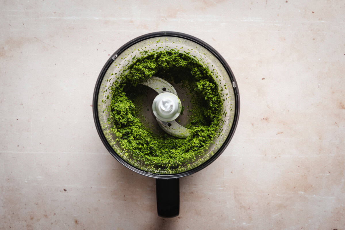 Top view of a food processor bowl containing pesto on a light beige surface. The blade is visible in the center, surrounded by the vibrant green mixture.