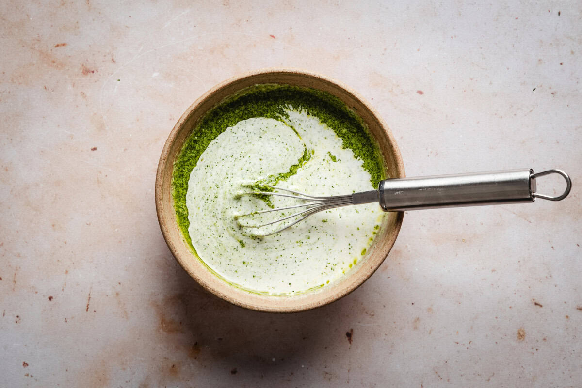 A bowl filled with pesto and cream mixture being gently combined. A small metal whisk rests inside the bowl. The surface below is light-colored and speckled.