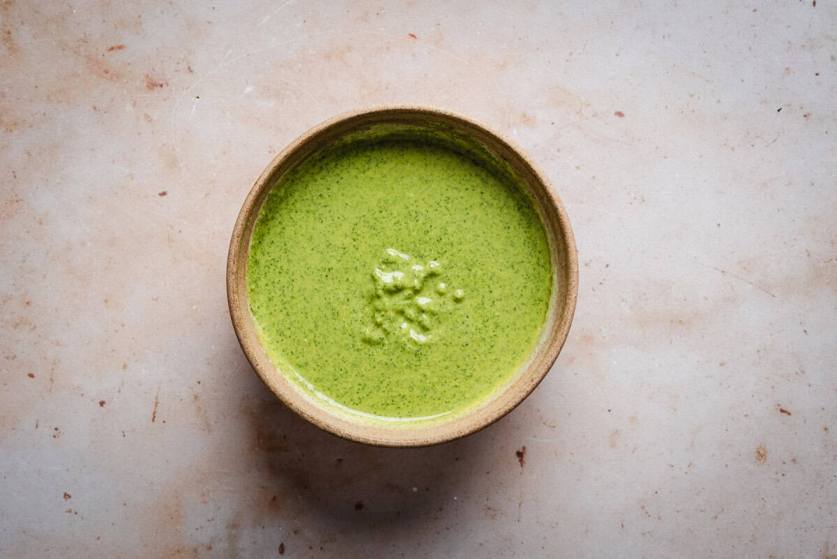 A bowl filled with a vibrant creamy pesto sauce on a light, speckled surface. The sauce appears creamy and textured, with small bubbles on its surface. The bowl is simple and round, complementing the earthy tones of the background.