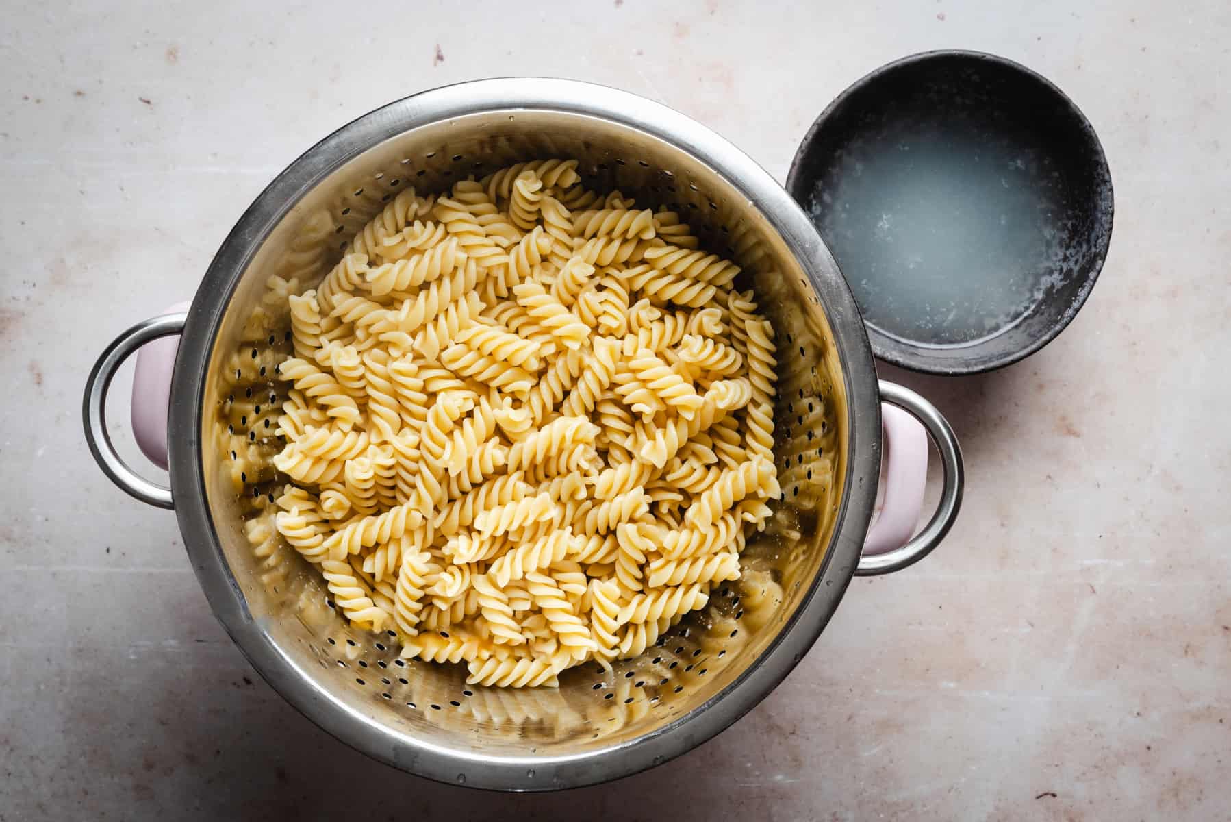 A metal colander filled with cooked fusilli pasta on a light countertop. Next to the colander is a dark, round bowl containing a small amount of cloudy water.