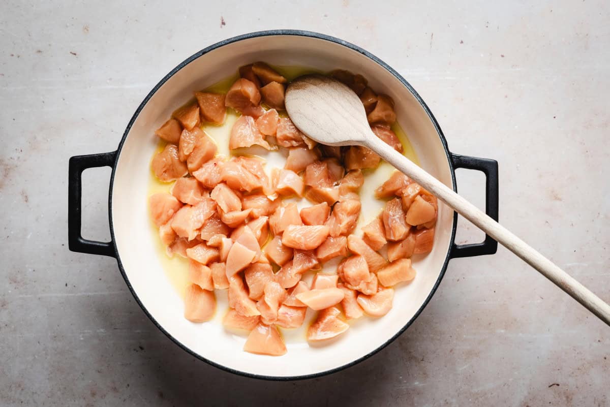 Top view of diced raw chicken in a white pot with a wooden spoon. The chicken pieces are surrounded by olive oil, ready for cooking. The pot has black handles and sits on a light-colored surface.