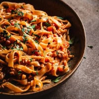 A rustic bowl of pork ragu and tagliatelle pasta garnished with fresh parsley. The dish is set on a textured surface, with a warm, inviting atmosphere.