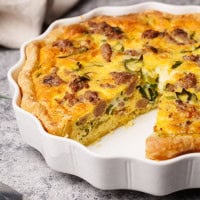 A close-up of a sausage quiche with a golden crust is shown in a round, fluted white dish. The quiche has browned sausage pieces and bits of green onion. A slice has been removed, revealing the filling. It's on a gray surface.