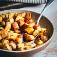 A bowl of golden-brown skillet potatoes, with a spoon resting inside. The potatoes are crispy and caramelized, served on a rustic ceramic plate. A striped cloth is partially visible in the background.