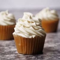 A vanilla cupcake with a swirl of stabilized whipped cream frosting on top, sitting on a textured surface, with two similar cupcakes blurred in the background.