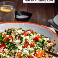 A bowl of colorful tabouli salad with cherry tomatoes, cucumber, parsley, and crumbled cheese on a table. A fork rests on the plate's edge. In the background, there's a glass of water and a stack of plates. Text at the top reads "Tabouli Salad, pinchandswirl.com.
