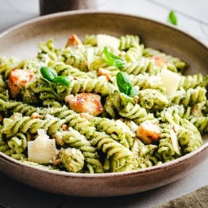 A shallow bowl of rotini pasta tossed in green pesto sauce, topped with grilled chicken pieces, shaved parmesan, pine nuts, and fresh basil leaves.