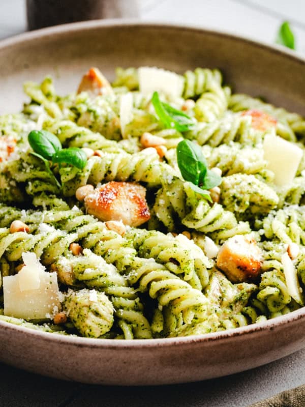 A shallow bowl of rotini pasta tossed in green pesto sauce, topped with grilled chicken pieces, shaved parmesan, pine nuts, and fresh basil leaves.