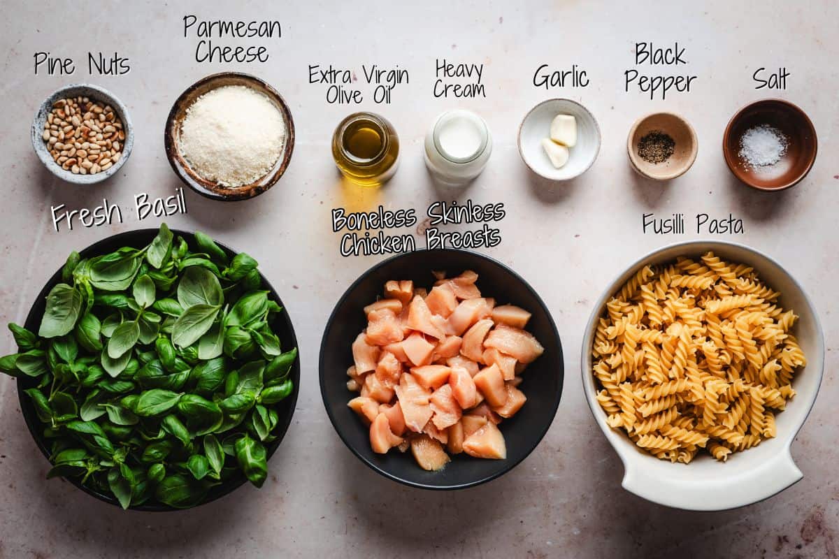 Ingredients for Chicken Pesto Pasta on a countertop: pine nuts, shredded parmesan cheese, extra virgin olive oil, heavy cream, chopped garlic, black pepper, salt, fresh basil leaves, diced boneless skinless chicken breasts, and uncooked fusilli pasta.