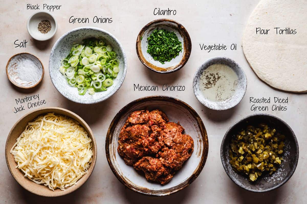 A top view of bowls containing ingredients for chorizo quesadillas including black pepper, sliced green onions, chopped cilantro, salt, shredded Monterey Jack cheese, Mexican chorizo, vegetable oil, roasted diced green chiles, and flour tortillas.