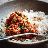 A close-up of a bowl filled with white rice and several mexican meatballs in chipotle sauce, garnished with chopped herbs, with a fork resting on the side of the bowl.