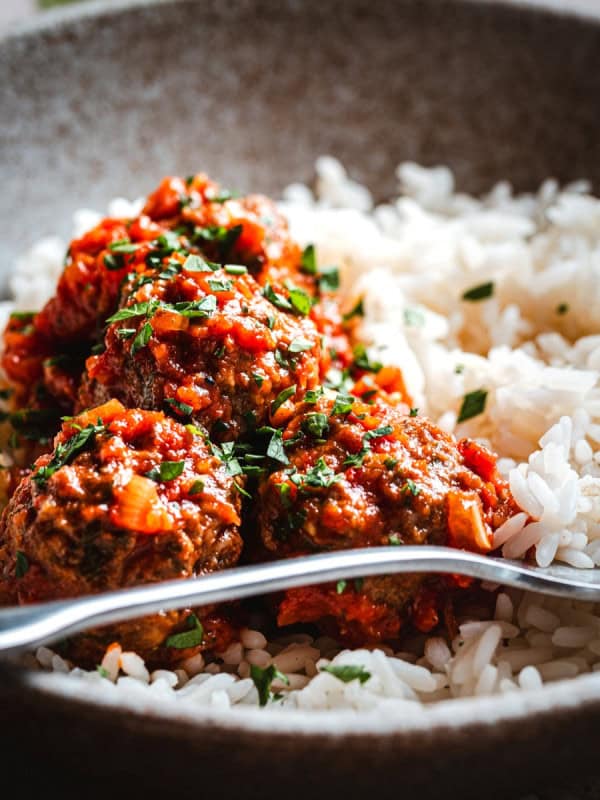 A close-up of a bowl filled with white rice and several mexican meatballs in chipotle sauce, garnished with chopped herbs, with a fork resting on the side of the bowl.