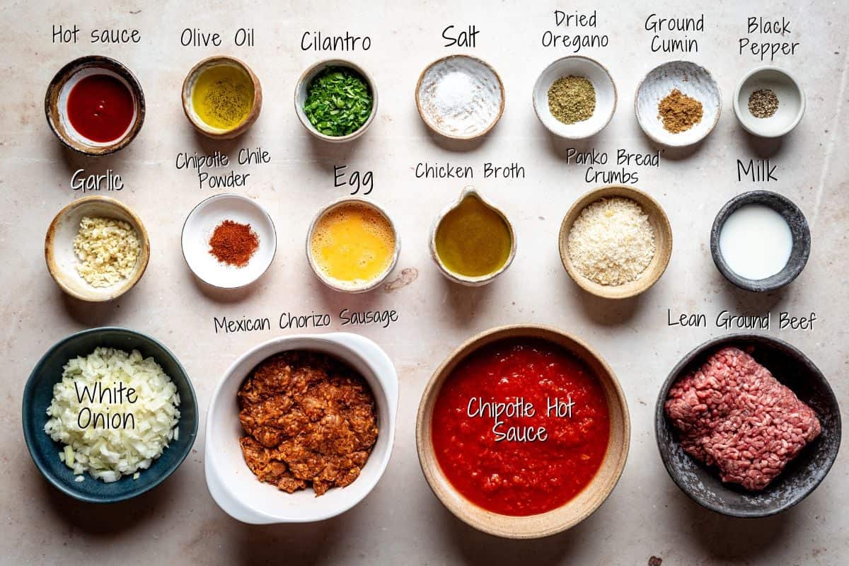 An overhead view of ingredients to make mexican meatballs and chipotle sauce in bowls, including hot sauce, olive oil, cilantro, salt, oregano, cumin, pepper, garlic, chipotle powder, egg, broth, breadcrumbs, milk, onion, chorizo, chipotle sauce, and ground beef.