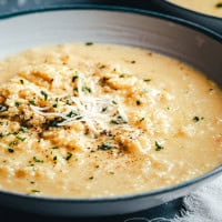 A close-up of a bowl of pastina topped with shredded cheese, chopped herbs, and freshly ground black pepper. The risotto looks rich and smooth, served in a shallow gray bowl.