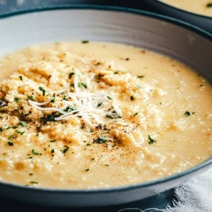 A close-up of a bowl of pastina topped with shredded cheese, chopped herbs, and freshly ground black pepper. The risotto looks rich and smooth, served in a shallow gray bowl.