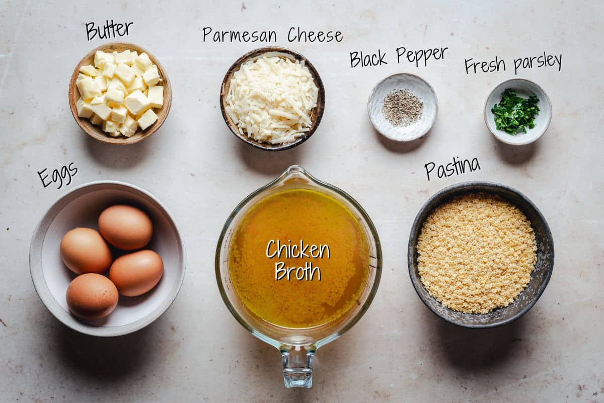 Overhead view of ingredients in bowls: cubed butter, shredded Parmesan cheese, black pepper, chopped fresh parsley, eggs, chicken broth in a measuring cup, and uncooked pastina pasta. Each item is labeled.