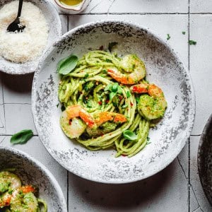 A rustic bowl of shrimp pesto pasta with pasta and pesto sauce, with shrimp, fresh basil, and red pepper flakes sits on a stone-patterned table next to a small dish of grated cheese.