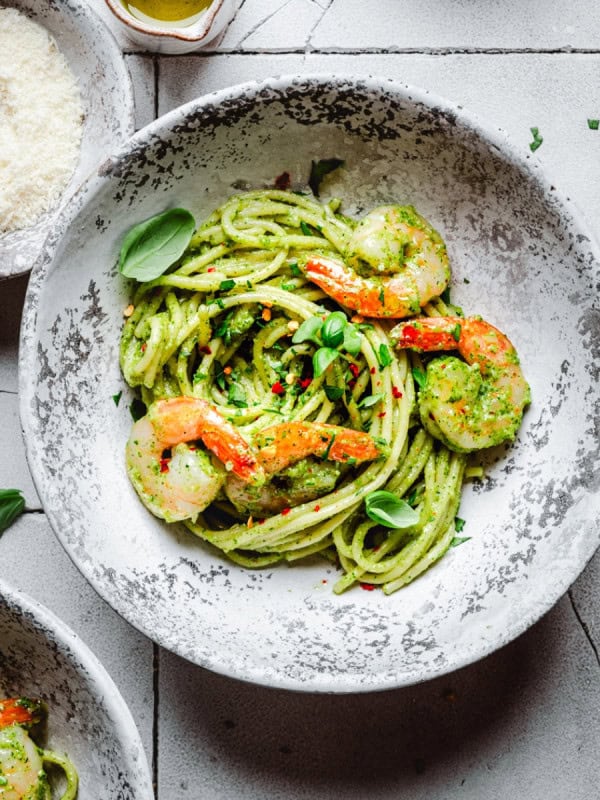 A rustic bowl of shrimp pesto pasta with pasta and pesto sauce, with shrimp, fresh basil, and red pepper flakes sits on a stone-patterned table next to a small dish of grated cheese.