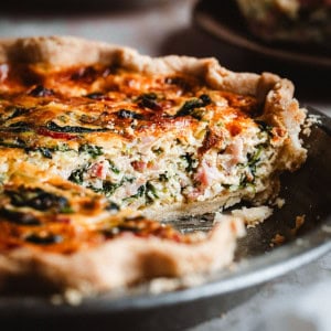A close-up of a freshly baked spinach bacon quiche in a metal pie dish, with a slice missing. The quiche has a golden crust and a filling with visible spinach, ham, and cheese.