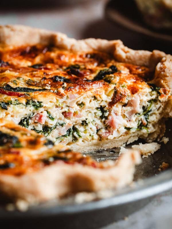 A close-up of a freshly baked spinach bacon quiche in a metal pie dish, with a slice missing. The quiche has a golden crust and a filling with visible spinach, ham, and cheese.