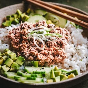 A bowl of white rice topped with creamy tuna, sliced avocado, cucumber, and green onions, with a pair of wooden chopsticks resting on the edge.
