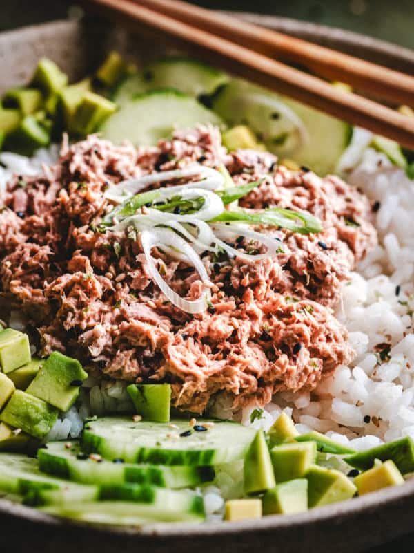 A bowl of white rice topped with creamy tuna, sliced avocado, cucumber, and green onions, with a pair of wooden chopsticks resting on the edge.