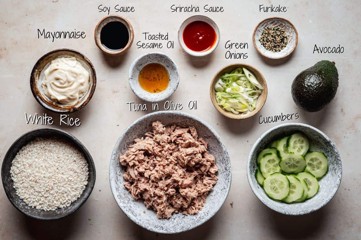 A variety of ingredients in bowls for a tuna rice bowl, including mayonnaise, soy sauce, sriracha, furikake, toasted sesame oil, green onions, avocado, cucumbers, white rice, and tuna in olive oil, all labeled.