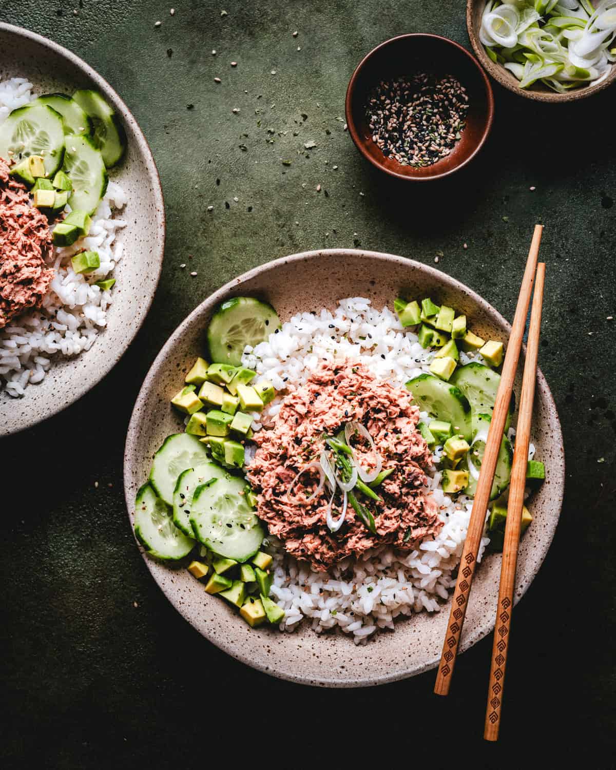 A bowl with white rice, sliced cucumbers, diced avocado, and a mound of creamy tuna, garnished with green onions. Chopsticks rest on the bowl. A small bowl of sesame seeds and sliced scallions are nearby.
