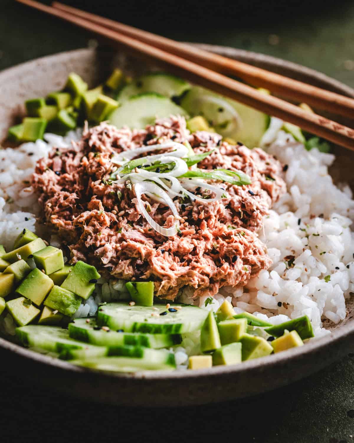 A bowl of white rice topped with creamy flaked tuna, sliced avocado, cucumber, and thinly sliced green onions, with wooden chopsticks resting on the bowl’s edge.