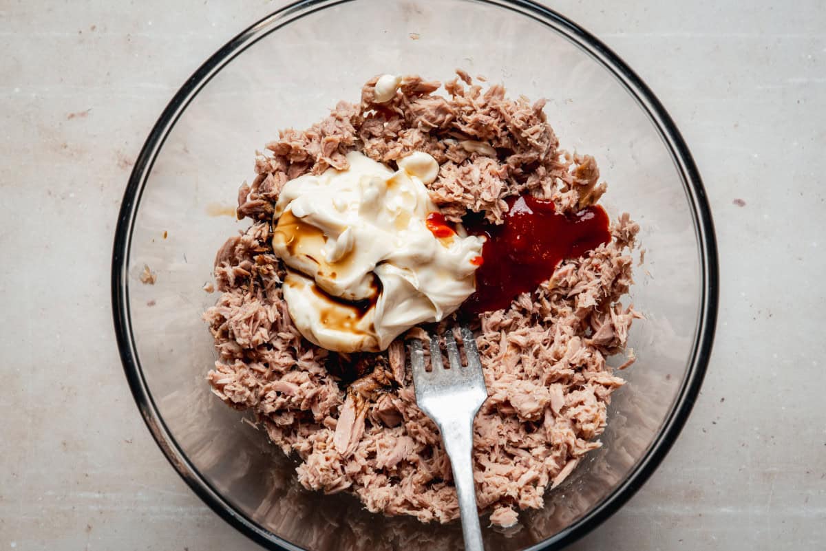 A glass bowl filled with shredded tuna, mayonnaise, sriracha sauce, and soy sauce, with a fork resting in the mixture, ready to be stirred.