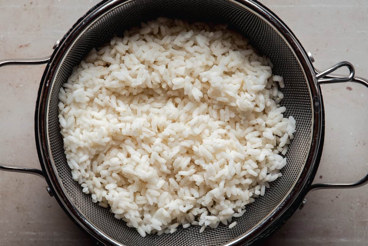 Cooked white rice sits in a metal mesh strainer placed on a light-colored surface, with the rice grains appearing separate and fluffy.