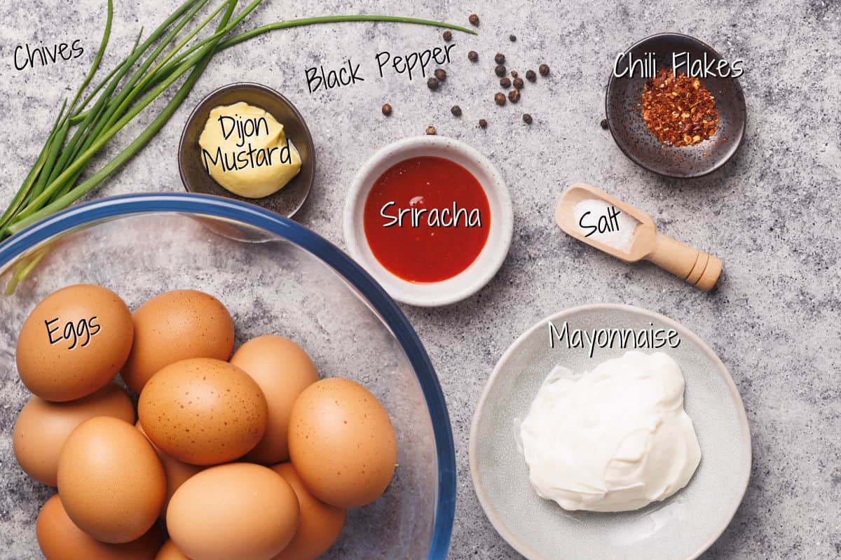 A top view of ingredients on a table: eggs in a bowl, mayonnaise on a plate, sriracha sauce, Dijon mustard, chives, black peppercorns, chili flakes, and salt in a scoop, each labeled with handwritten text.